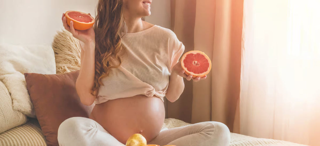 Eine Frau in fortgeschrittener Schwangerschaft sitzt mit angezogenen Beinen auf einem Sofa und hält eine halbierte Grapefruit in der Hand.