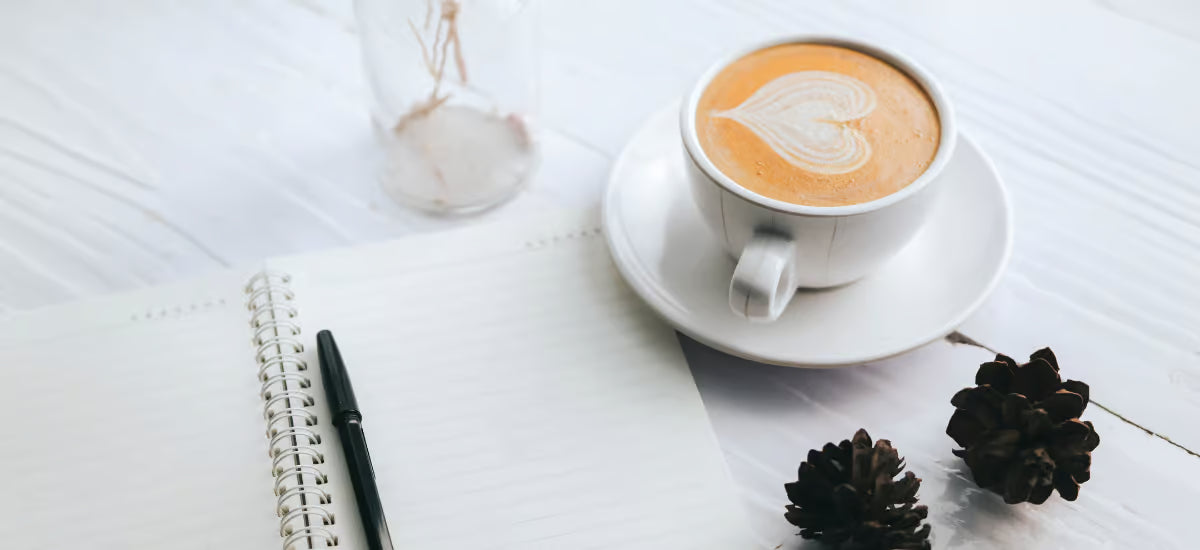 Eine Tasse Kaffee und ein Notizbuch mit einem Stift auf einem weißen Tisch mit zwei Tannenzapfen.