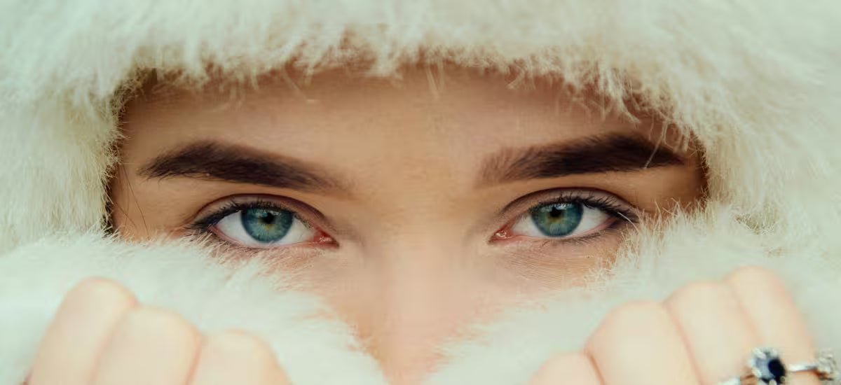 Nahaufnahme einer Frau mit blauen Augen, eingehüllt in weiches weißes Fell, die ihr Gesicht vor winterlicher Kälte schützt.