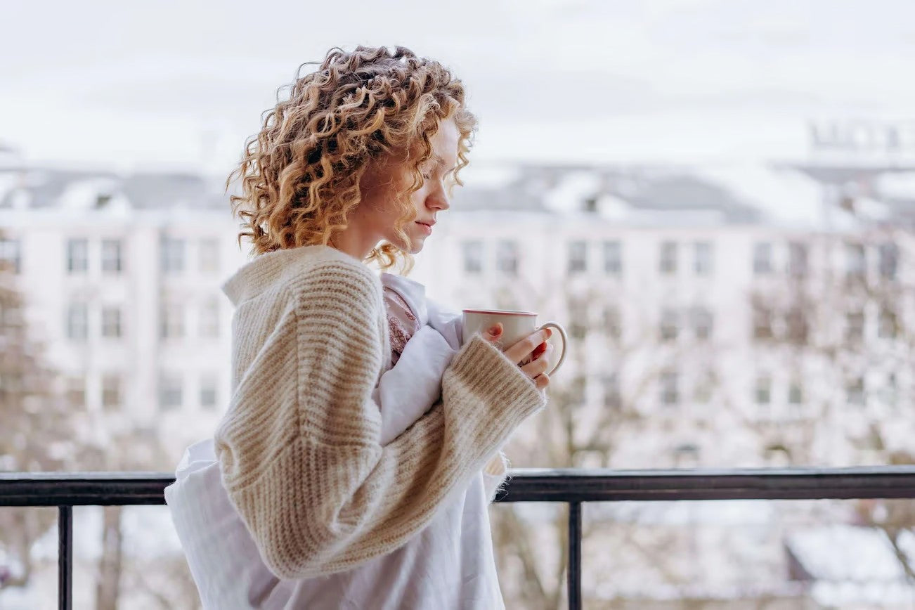 Junge Frau mit lockigen Haaren steht auf dem Balkon und hält eine Tasse in den Händen