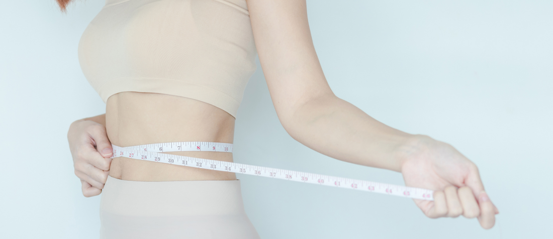 Frau in beiger Sportkleidung misst ihre Taille mit einem Maßband