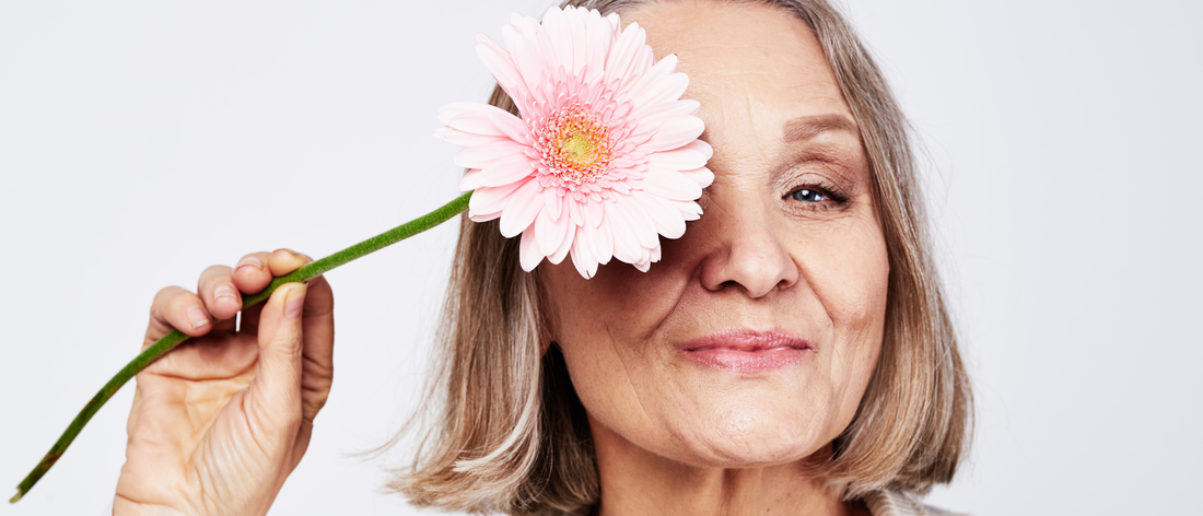 Lächelnde Frau in den Wechseljahren mit strahlender Haut und schönen Haaren nach der Einnahme von Kollagen-Trinkampullen, die eine rosa Blume vor ihr Auge hält.
