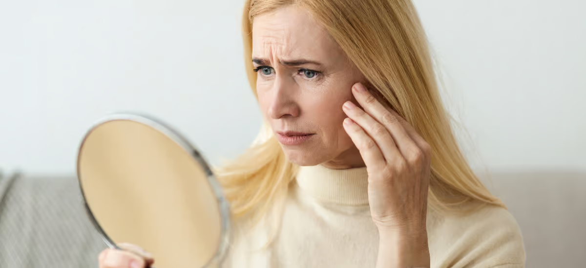 Eine Frau betrachtet ihr Gesicht im Spiegel und runzelt die Stirn|||Primabiotic Collagen