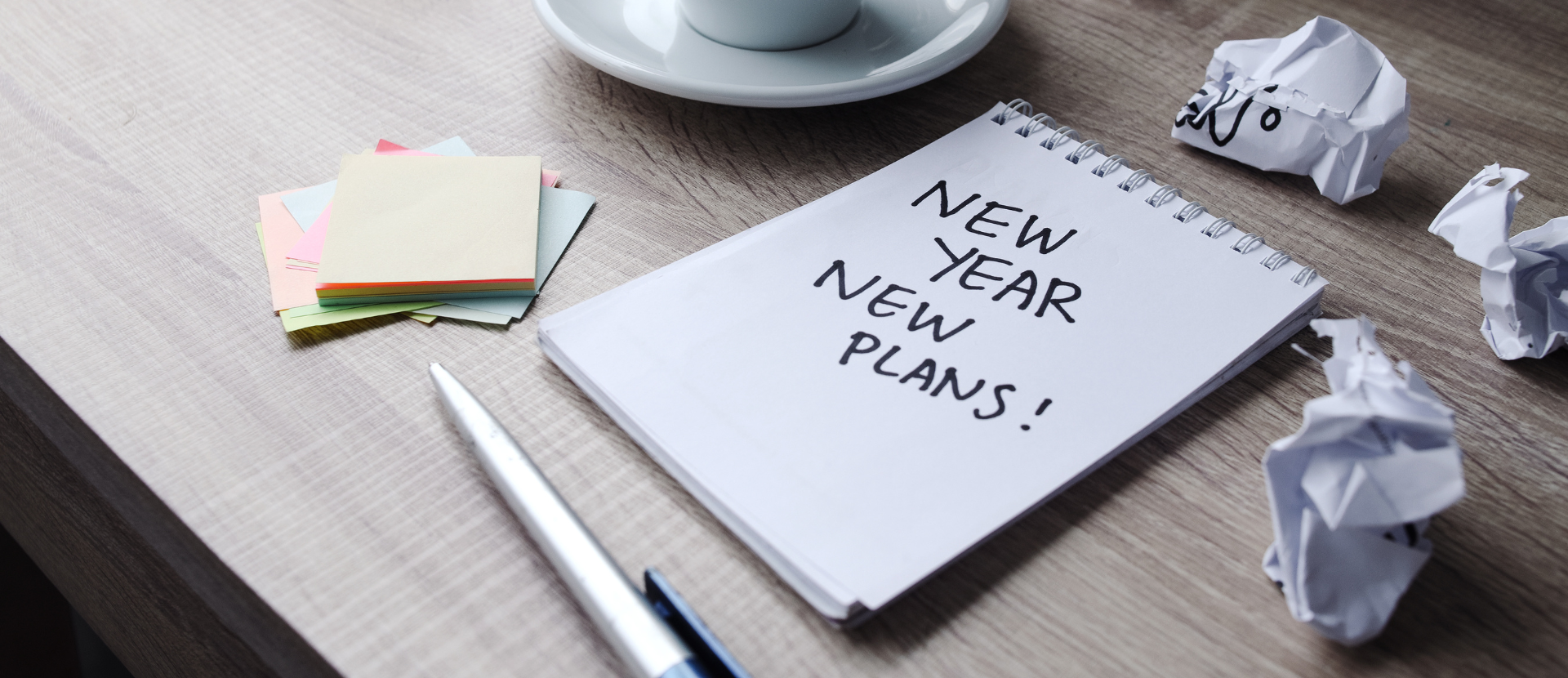 Notizbuch mit der Aufschrift „New Year New Plans“ auf Schreibtisch neben Stift und Haftnotiz