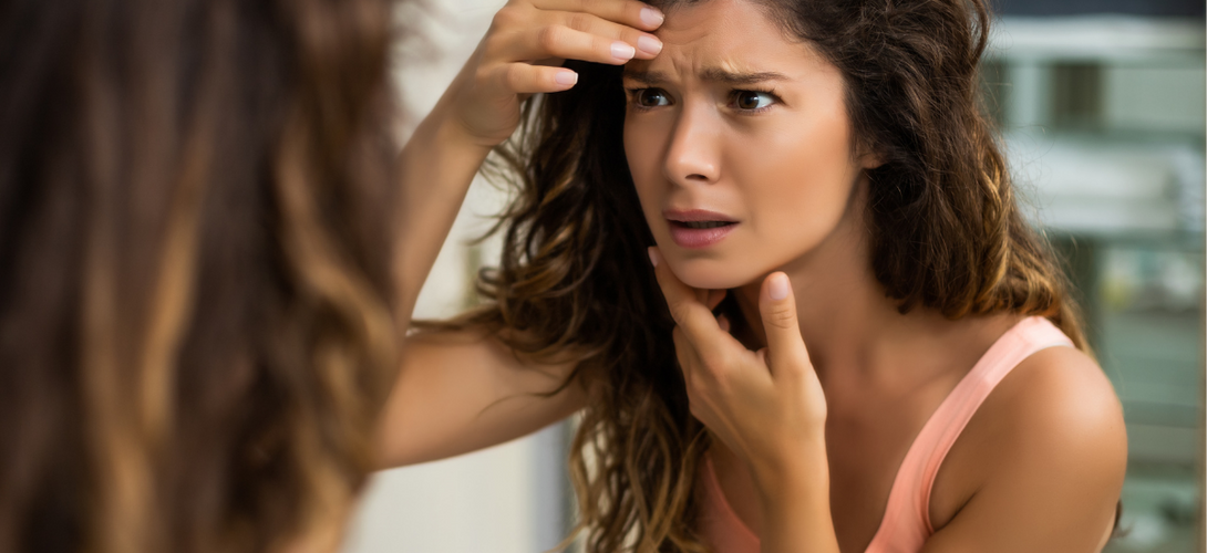 Frau mit besorgtem Blick betrachtet ihr Gesicht im Spiegel und bemerkt Hautunreinheiten