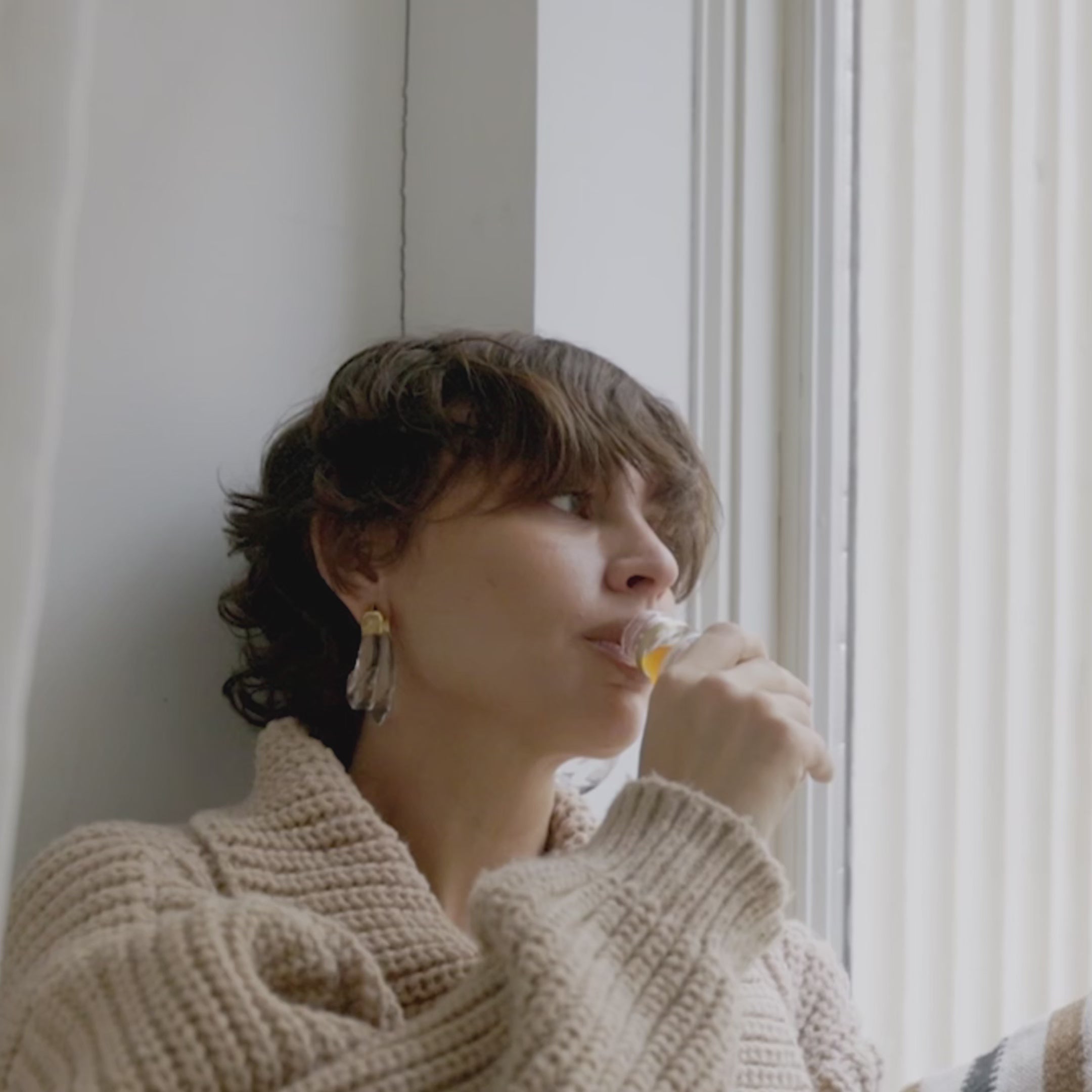 Frau mit kurzem Haar und beigen Strickpullover trinkt am Fenster einen Shot Primabiotic Hyaluron.