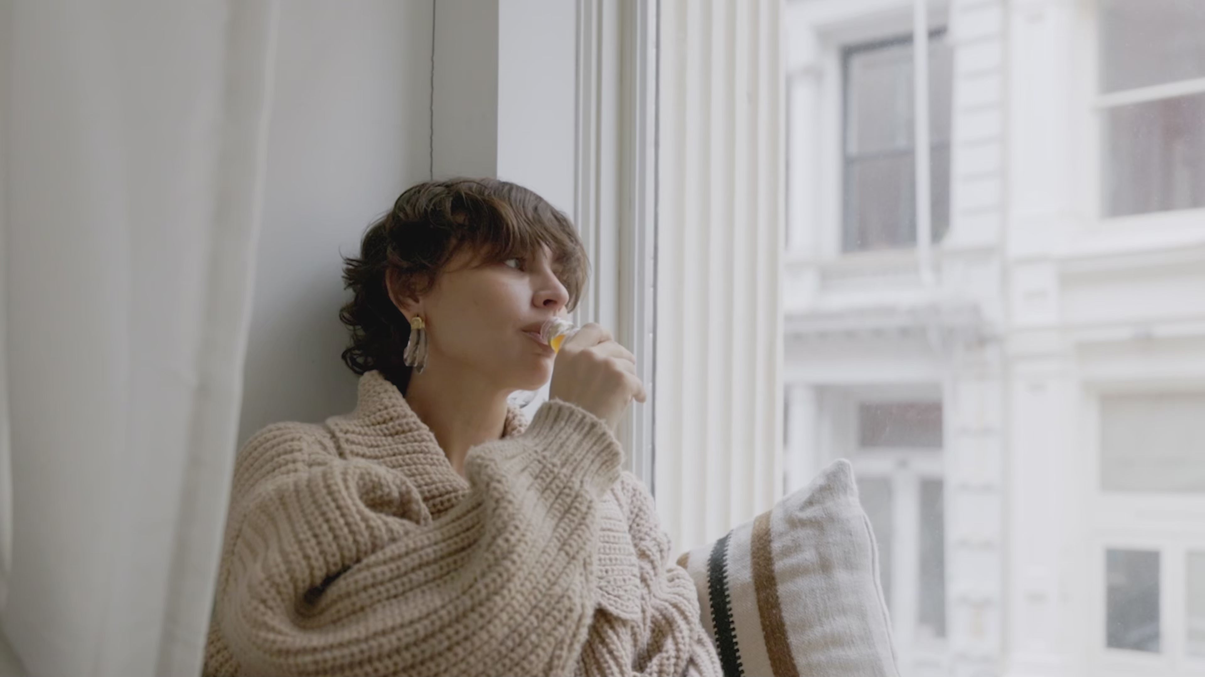 Frau mit kurzem Haar und beigen Strickpullover trinkt am Fenster einen Shot Primabiotic Hyaluron.